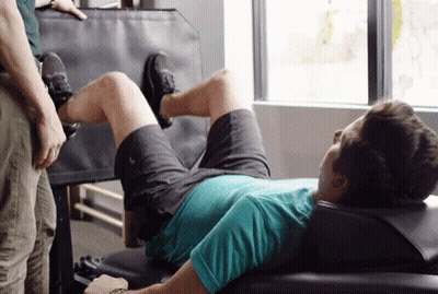 A Man Is Laying On A Bench In A Gym While A Man Stands Behind Him . GIF