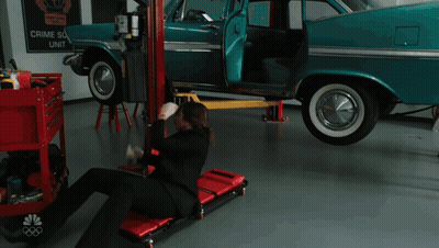 A Man Is Laying On A Rolling Chair In Front Of A Car That Has A Sign That Says Crime Scene Unit On It GIF