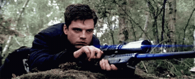 A Man Is Laying On The Ground Holding A Sniper Rifle In A Forest . GIF