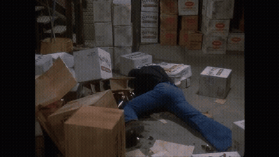 A Man Is Laying On The Ground In A Warehouse Surrounded By Boxes Including One That Says Seven GIF