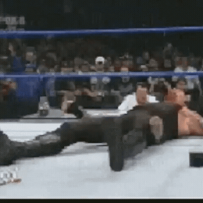 A Man Is Laying On The Ground In A Wrestling Ring While A Crowd Watches . GIF