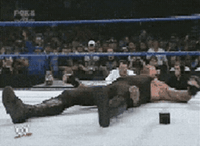 A Man Is Laying On The Ground In A Wrestling Ring While A Crowd Watches . GIF
