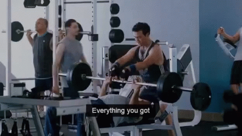 A Man Is Lifting A Barbell In A Gym While Another Man Watches . GIF