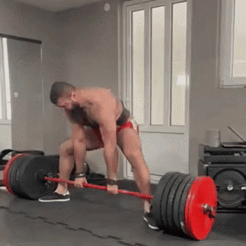 A Man Is Lifting A Barbell In A Gym While Wearing Shorts . GIF