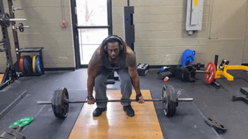 A Man Is Lifting A Barbell In A Gym With Headphones On GIF
