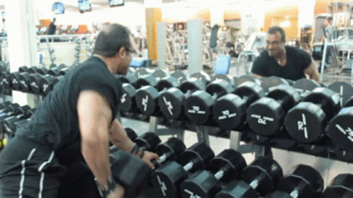A Man Is Lifting A Dumbbell In Front Of A Row Of Dumbbells With The Letters Dc On Them GIF