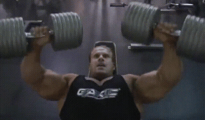 A Man Is Lifting Dumbbells In A Gym Wearing A Black Tank Top That Says Fake GIF