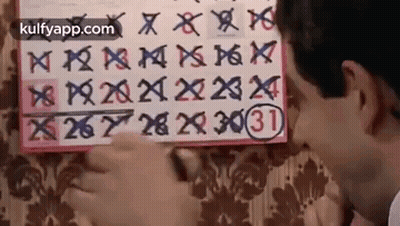 A Man Is Looking At A Calendar On A Wall And Writing On It . GIF