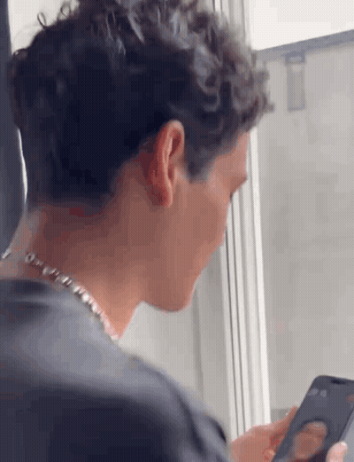 A Man Is Looking At A Cell Phone While Sitting In Front Of A Window . GIF