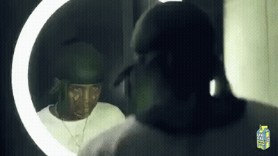 A Man Is Looking At Himself In A Mirror With A Carton Of Lychee Juice Behind Him . GIF