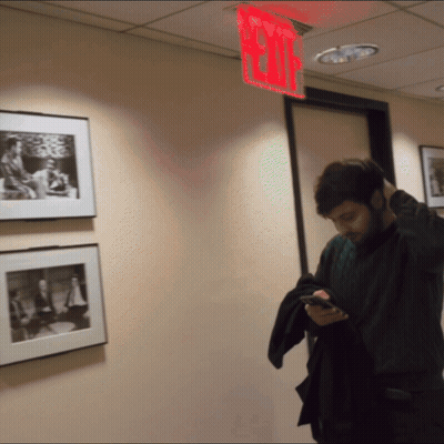 A Man Is Looking At His Phone In A Hallway With A Red Exit Sign Above Him GIF