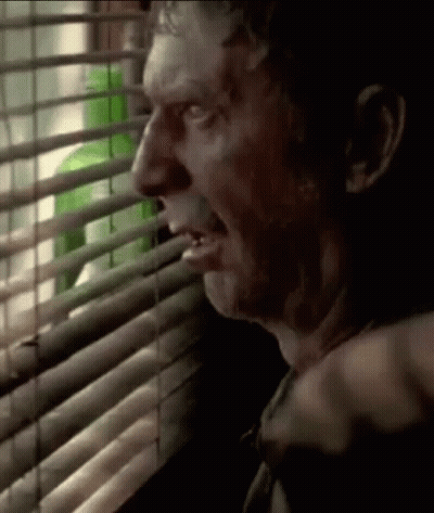 A Man Is Looking Out A Window With Blinds And A Green Bottle Behind Him . GIF
