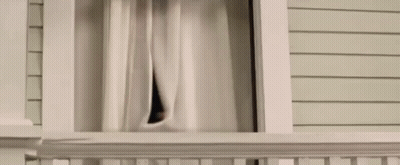 A Man Is Looking Out Of A Window With Curtains . GIF
