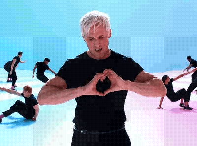 A Man Is Making A Heart Shape With His Hands While A Group Of People Are Dancing In The Background . GIF