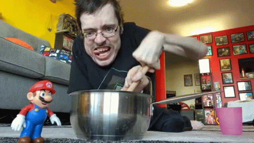 A Man Is Mixing Something In A Bowl With A Mario Toy In The Foreground GIF