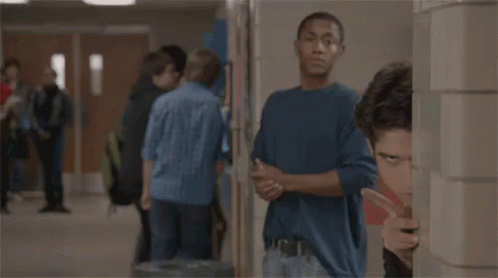 A Man Is Peeking Out From Behind A Wall In A Hallway While A Group Of People Are Standing In The Background . GIF