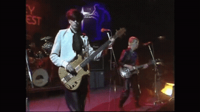 A Man Is Playing A Bass Guitar On A Stage While Another Man Sings Into A Microphone . GIF