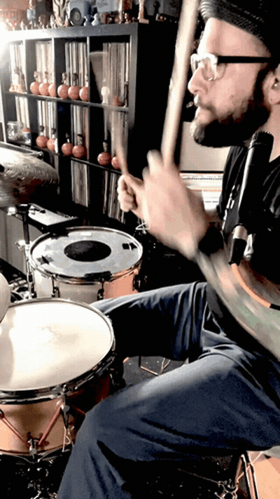 A Man Is Playing A Drum Set In Front Of A Shelf With Records On It GIF