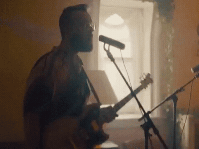 A Man Is Playing A Guitar And Singing Into A Microphone While Standing In Front Of A Window . GIF