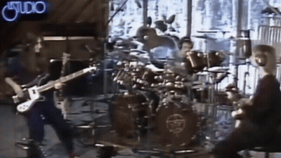 A Man Is Playing A Guitar In Front Of A Drum Set With A Neon Sign That Says Studio GIF