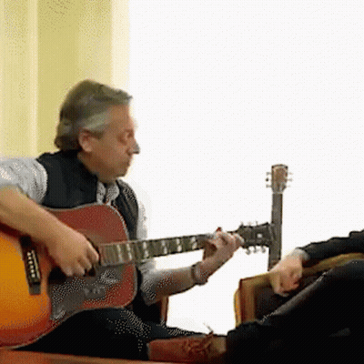 A Man Is Playing An Acoustic Guitar While Another Man Sits In A Chair GIF
