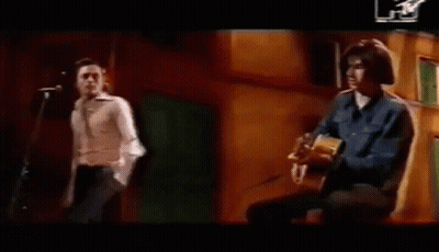 A Man Is Playing An Acoustic Guitar While Another Man Stands Behind Him . GIF