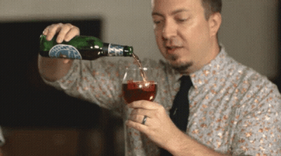 A Man Is Pouring A Drink From A Bottle Into A Glass With A Blue Label That Says ' A ' On It GIF