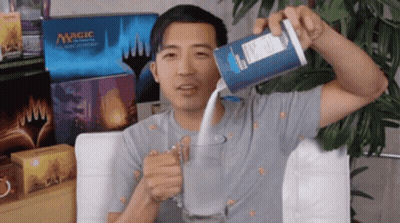 A Man Is Pouring Milk Into A Glass In Front Of Boxes Of Magic The Gathering Cards GIF