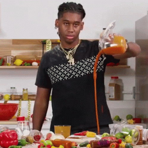 A Man Is Pouring Orange Juice Into A Glass While Standing In A Kitchen Surrounded By Vegetables . GIF