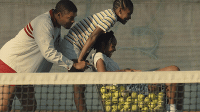 A Man Is Pushing A Woman On A Tennis Court With A Basket Full Of Tennis Balls GIF