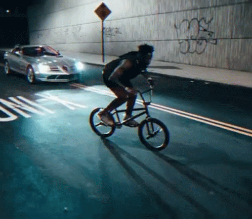 A Man Is Riding A Bike Down A Street With Graffiti On The Wall Behind Him GIF