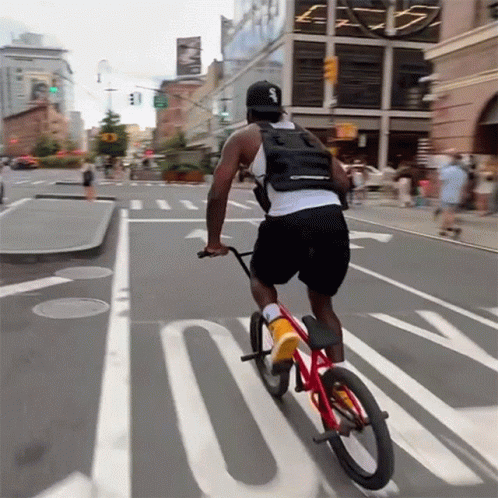 A Man Is Riding A Bike Down A Street With The Letter S On His Back GIF