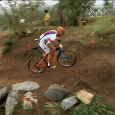 A Man Is Riding A Bike On A Dirt Trail With The Olympic Rings In The Background GIF