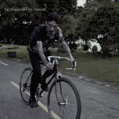 A Man Is Riding A Bike On A Road With La Guarimba Film Festival Written On The Bottom Right GIF