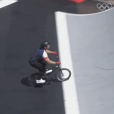 A Man Is Riding A Bike On A Road With The Olympic Rings In The Background GIF