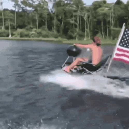 A Man Is Riding A Boat In The Water With An American Flag Behind Him . GIF