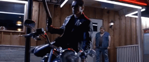A Man Is Riding A Motorcycle In Front Of A Building While Another Man Stands Behind Him . GIF