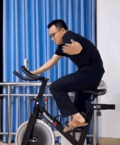 A Man Is Riding A Reebok Exercise Bike In Front Of A Blue Curtain GIF