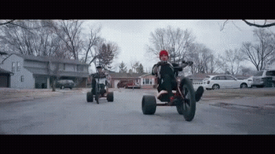 A Man Is Riding A Tricycle Down A Street Next To A Man Riding A Bike . GIF