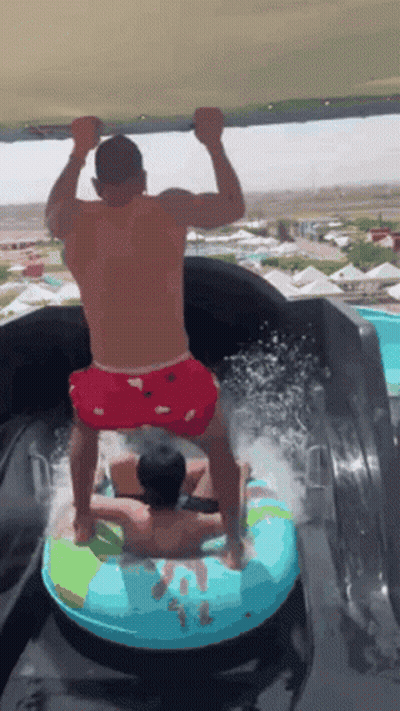A Man Is Riding A Tube Down A Water Slide While Another Man Stands On Top Of It . GIF