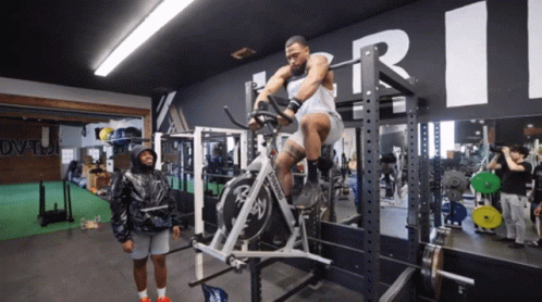 A Man Is Riding An Exercise Bike In A Gym With A Large R On The Wall Above Him GIF