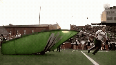 A Man Is Running On A Football Field With A Large Green Flag That Says ' M ' On It GIF