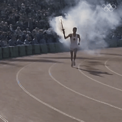 A Man Is Running With A Torch On A Track With The Olympic Rings In The Background GIF