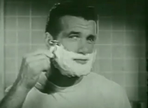 A Man Is Shaving His Face In A Bathroom With A Razor . GIF