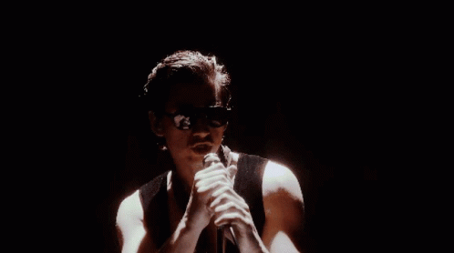 A Man Is Singing Into A Microphone In A Dark Room While Wearing Sunglasses . GIF