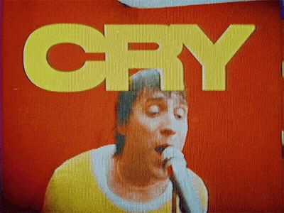 A Man Is Singing Into A Microphone In Front Of A Red Wall That Says Cry GIF