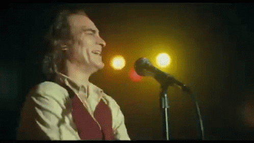 A Man Is Singing Into A Microphone On A Stage In A Dark Room . GIF