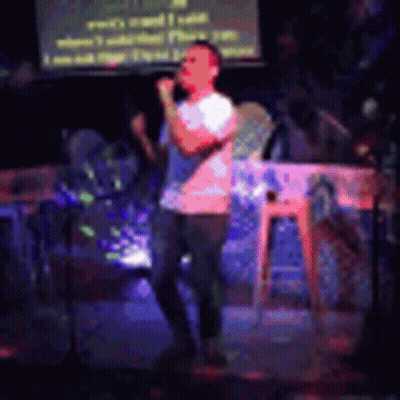 A Man Is Singing Into A Microphone On A Stage In Front Of A Screen . GIF