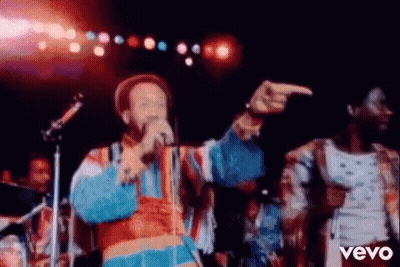 A Man Is Singing Into A Microphone On A Stage While Another Man Points At Him . GIF