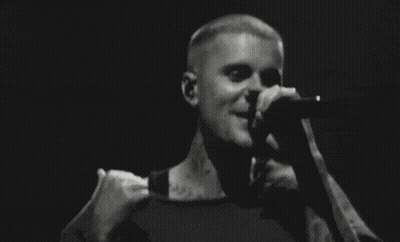 A Man Is Singing Into A Microphone On Stage In A Black And White Photo . GIF